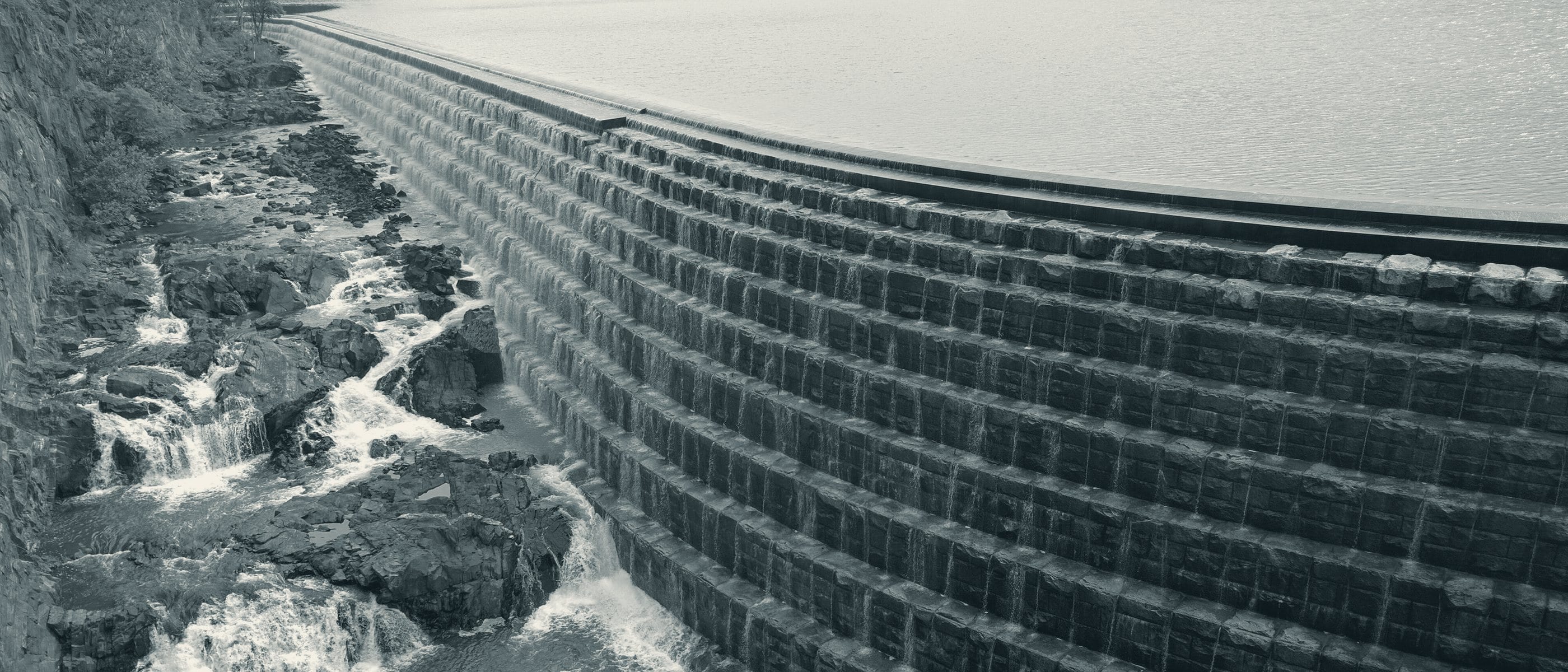 The stepped stone spillway of the New Croton Dam, water falling evenly down each course beside the rocks of the gorge.